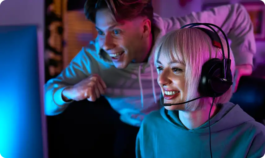Jonge gamer met een headset op, glimlachend terwijl haar teamgenoot achter haar feestviert in een levendige gamingomgeving.